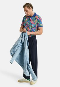 Blaues, pinkes und grünes Polohemd mit Blumenmuster, getragen mit dunklen Hosen. Hält eine hellblaue Jeansjacke. Weiße Sneaker mit grünen Akzenten.