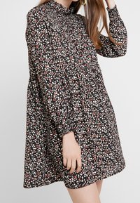 Mujer vestida con un vestido negro de manga larga con pequeño estampado floral en rosa y blanco, con yugo fruncido y puños elásticos, de pie frente a un fondo liso.