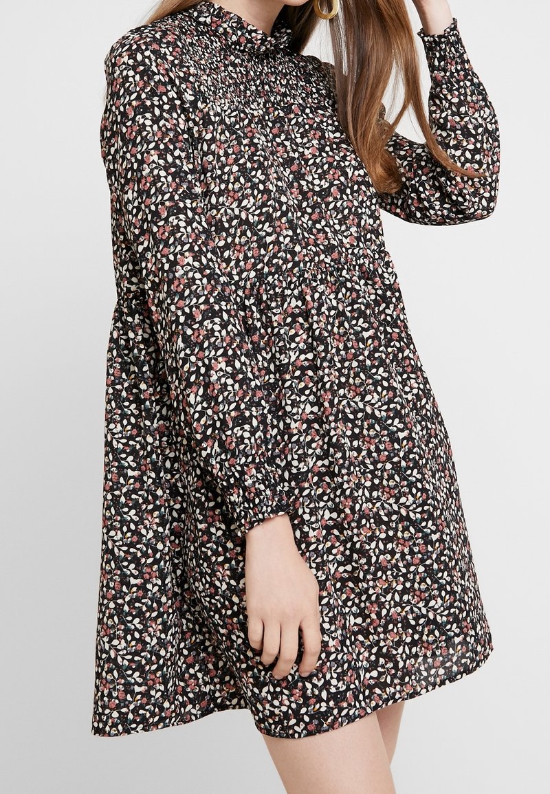 Mujer vestida con un vestido negro de manga larga con pequeño estampado floral en rosa y blanco, con yugo fruncido y puños elásticos, de pie frente a un fondo liso.