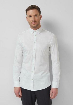 Junger Mann mit einem weißen, gemusterten Hemd mit langen Ärmeln und Knöpfen sowie schwarzer Hose, der vor einem schlichten hellgrauen Hintergrund steht.