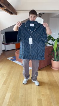 Camisa de manga curta preta e azul com padrão geométrico, detalhes brancos e um bolso frontal. Etiqueta anexada, exibida por um modelo.