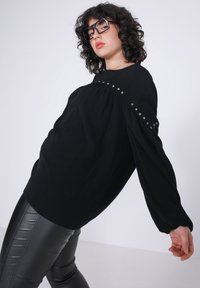 Zwarte top met lange mouwen, ronde halslijn, gerimpeld schouderontwerp en zilveren studs langs de naad. Gestyled met zwarte glanzende broeken.