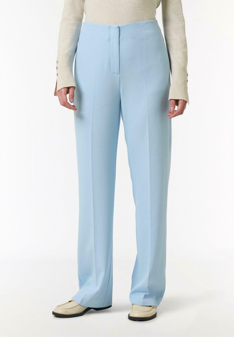 Personne portant un pantalon large bleu clair, un haut beige à manches longues et des mocassins crème sur fond blanc.
