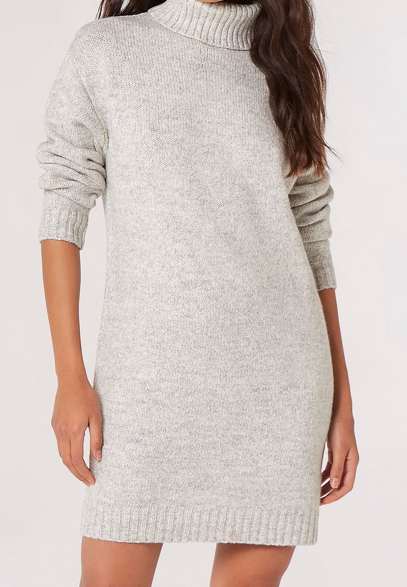 Femme portant une robe-pull en tricot gris clair, à manches longues, avec un col roulé haut et des poignets et ourlet côtelés.