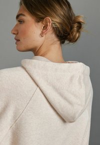 Hoodie beige clair avec une texture douce, doté d'une capuche, d'une coupe ample et de détails de couture discrets. Accentuée par de petites boucles d'oreilles.