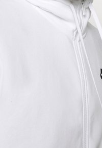 Vit zip-up hoodie gjord av mjukt tyg, med hög krage, snörning och en liten svart logotyp på bröstet.