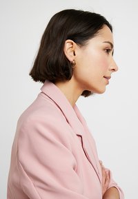 Blush-pinker Blazer mit Reverskragen, strukturierter Stoff und Dreiviertelärmeln; Person trägt goldene Creolen und hat kurzes dunkles Haar.