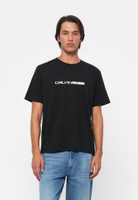 Black cotton t-shirt with short sleeves featuring a white "CALVIN KLEIN" logo across the chest. Casual fit, minimalistic design, round neckline.