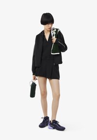 Conjunto atlético negro que incluye una chaqueta con cremallera y pantalones cortos con pliegues. Sosteniendo una botella de agua verde y usando zapatillas Lacoste moradas.
