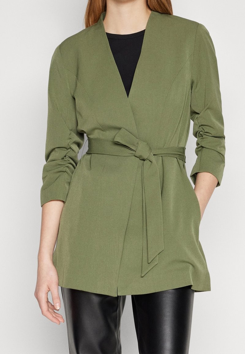 Femme portant un blazer ceinturé vert olive avec des manches froncées sur un top noir et un pantalon en cuir noir, debout avec une main dans la poche.