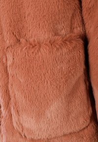 Faux fur frakke i blød, tekstureret ferskenfarve. Har en stor klaplomme med synlige sømme langs kanterne og et blødt udseende.