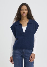 Veste tricotée marine à col en V profond et manches courtes, avec un ourlet côtelé. Portée sur une chemise blanche à manches longues et associée à un jean bleu.