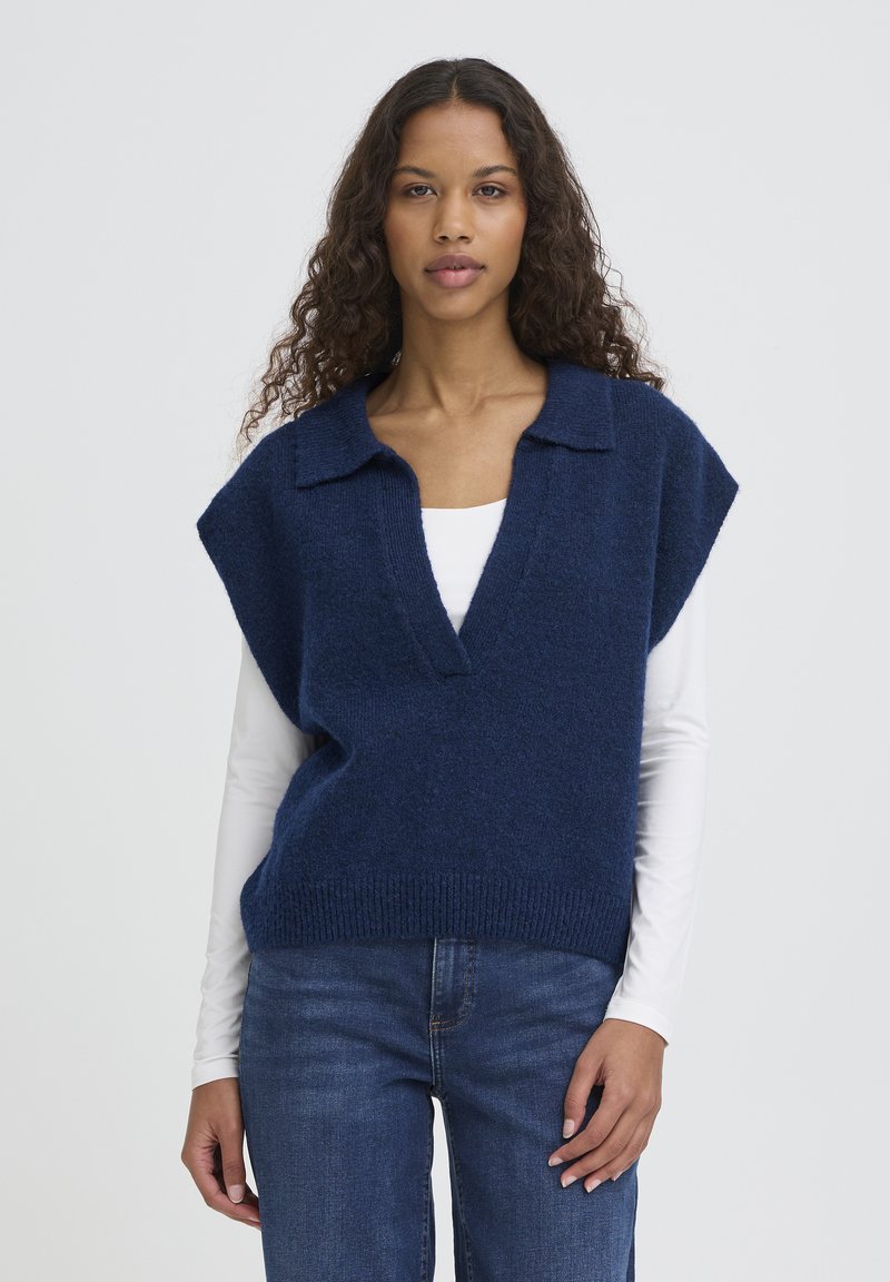 Veste tricotée marine à col en V profond et manches courtes, avec un ourlet côtelé. Portée sur une chemise blanche à manches longues et associée à un jean bleu.