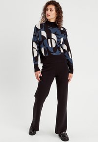 Maglione lavorato a maglia con collo alto, caratterizzato da un motivo geometrico in blu, nero e bianco. Abbinato a pantaloni svasati neri e scarpe nere.