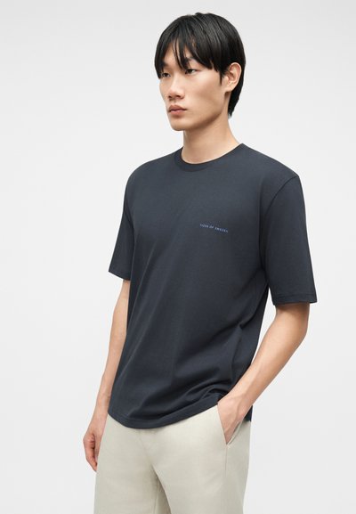 Νεαρός άνδρας που φοράει ένα σκουρόγκρι μπλουζάκι Tiger of Sweden και ανοιχτό μπεζ παντελόνι, στέκεται με το ένα χέρι στην τσέπη μπροστά σε απλό φόντο.