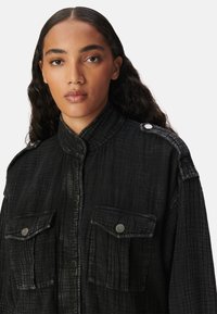 Camicia in tessuto nero con texture, colletto alto, due tasche frontali, bottoni a pressione e spalline sulle spalle. Finitura liscia.