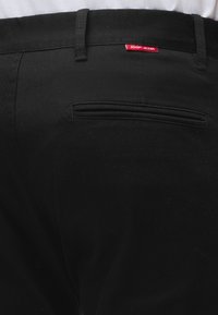 Schwarze Hose mit einer Gesäßtasche mit Paspel und einem kleinen roten "JOOP! JEANS" Etikett über der Tasche auf weißem Hemdhintergrund.