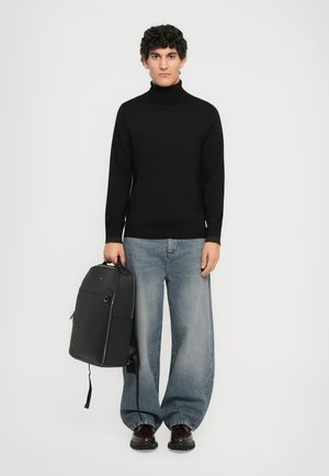 Man in black turtleneck, wide-leg faded jeans, brown shoes holding black backpack against plain white background.