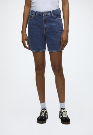 Άτομο που φοράει σορτς από τζιν σε μεσαίο μπλε χρώμα, λευκές κάλτσες, μαύρα αθλητικά παπούτσια και λευκό τοπ, στέκεται μπροστά σε ένα απλό λευκό φόντο.