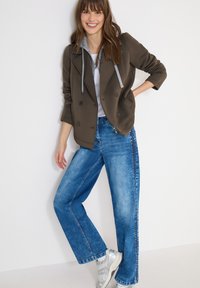 Femme souriante portant une veste marron à double boutonnage avec des détails de sweat à capuche gris, un jean large bleu et des baskets blanches, debout contre un mur blanc.