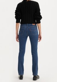 Jeans de mezclilla azul con diseño de pierna recta, lavado medio y un sutil patrón de costura en los bolsillos traseros. Combinados con un suéter negro de punto trenzado.