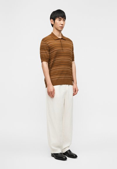 Άνδρας στέκεται φορώντας καφέ πουκαμίσα πόλο με μανίκια κοντά και σχέδια, λευκό παντελόνι με φαρδύ τελείωμα και μαύρα επίσημα παπούτσια, μπροστά σε απλό λευκό φόντο.