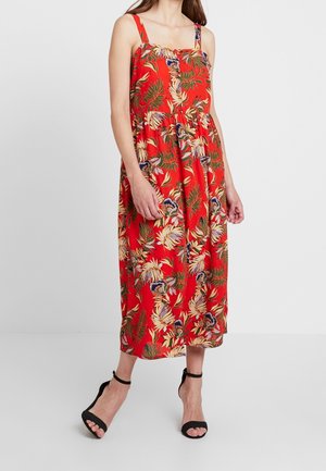 Vrouw draagt een rode mouwloze midijurk met tropische bladeren- en bloemenprint, gecombineerd met zwarte hoge hakken met enkelbandje tegen een witte achtergrond.