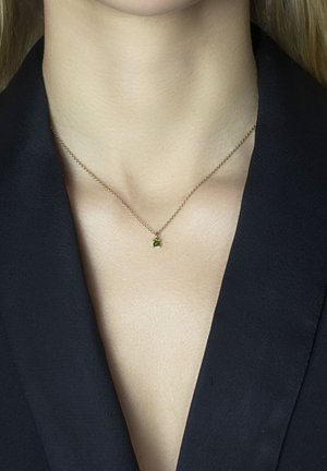 Goldene Halskette mit einem kleinen grünen Edelstein-Anhänger, der am Schlüsselbein getragen wird, im Kontrast zu einem dunklen Blazer. Schlichtes und elegantes Design.