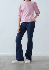 Světlounce růžová mikina s dlouhým rukávem a nafouklými rukávy, doplněná tmavě modrými zvonovými džíny a bílými teniskami, na neutrálním pozadí.