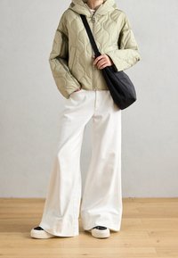 Veste matelassée vert olive avec capuche et fermeture éclair devant, associée à un pantalon blanc à jambes larges et un sac bandoulière noir. Baskets avec des accents noirs et blancs.