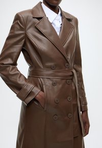 Gabardina de cuero marrón con botones cruzados, solapa con muesca, cintura con cinturón y bolsillos laterales. Textura suave, diseño entallado.