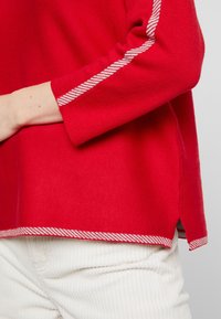 Pull en maille rouge avec une texture côtelée, comportant des accents à rayures blanches le long des manches et une fente latérale à l'ourlet.