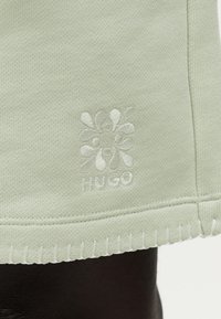 Ανοιχτοπράσινο πλεκτό ύφασμα με κεντητό αφηρημένο λουλούδι και κείμενο "HUGO" κοντά στην ραμμένη άκρη του τελειώματος.