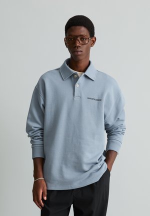 Jeune homme aux cheveux courts et portant des lunettes, vêtu d'un polo bleu clair à manches longues et d'un pantalon noir, debout devant un fond gris uni.