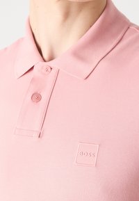 Chemise polo rose en tissu texturé, avec un col, deux boutons, et un patch logo carré étiqueté "BOSS" sur la poitrine.