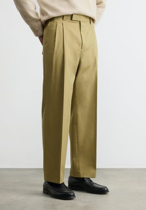 Hombre con pantalones plisados beige y mocasines de cuero negro, de pie sobre un suelo gris con fondo blanco.