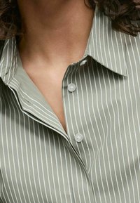 Light olive green shirt featuring vertical white stripes, a pointed collar, and two button closures on the neckline. Smooth fabric texture.