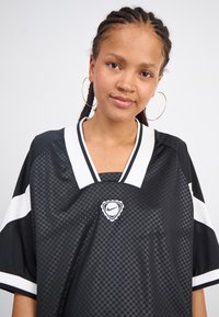 Black mesh shirt with a checkered pattern, white accents, and a v-neckline. Features a logo at the center and short, wide sleeves.