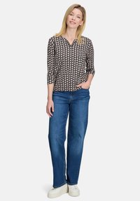 Drukowana bluzka z dekoltem w serek, rękawami 3/4 oraz geometrycznym wzorem w czerni i kremie, zestawiona z niebieskimi spodniami typu wide-leg i białymi butami.