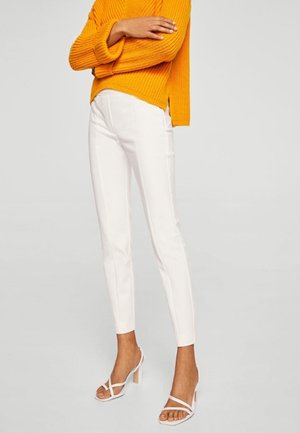 Osoba ubrana w białe obcisłe spodnie, białe sandały na wysokim obcasie z paskami oraz pomarańczowy prążkowany sweter z założonymi skrzyżowanymi ramionami.