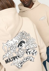 Sweat à capuche beige avec poche frontale et un grand imprimé graphique mettant en avant des designs abstraits et du texte en noir, présentant différentes textures et formes.