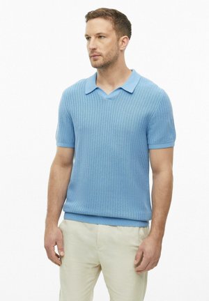 Man draagt een lichtblauw gebreid polohemd met korte mouwen en een beige broek, staand tegen een effen witte achtergrond.
