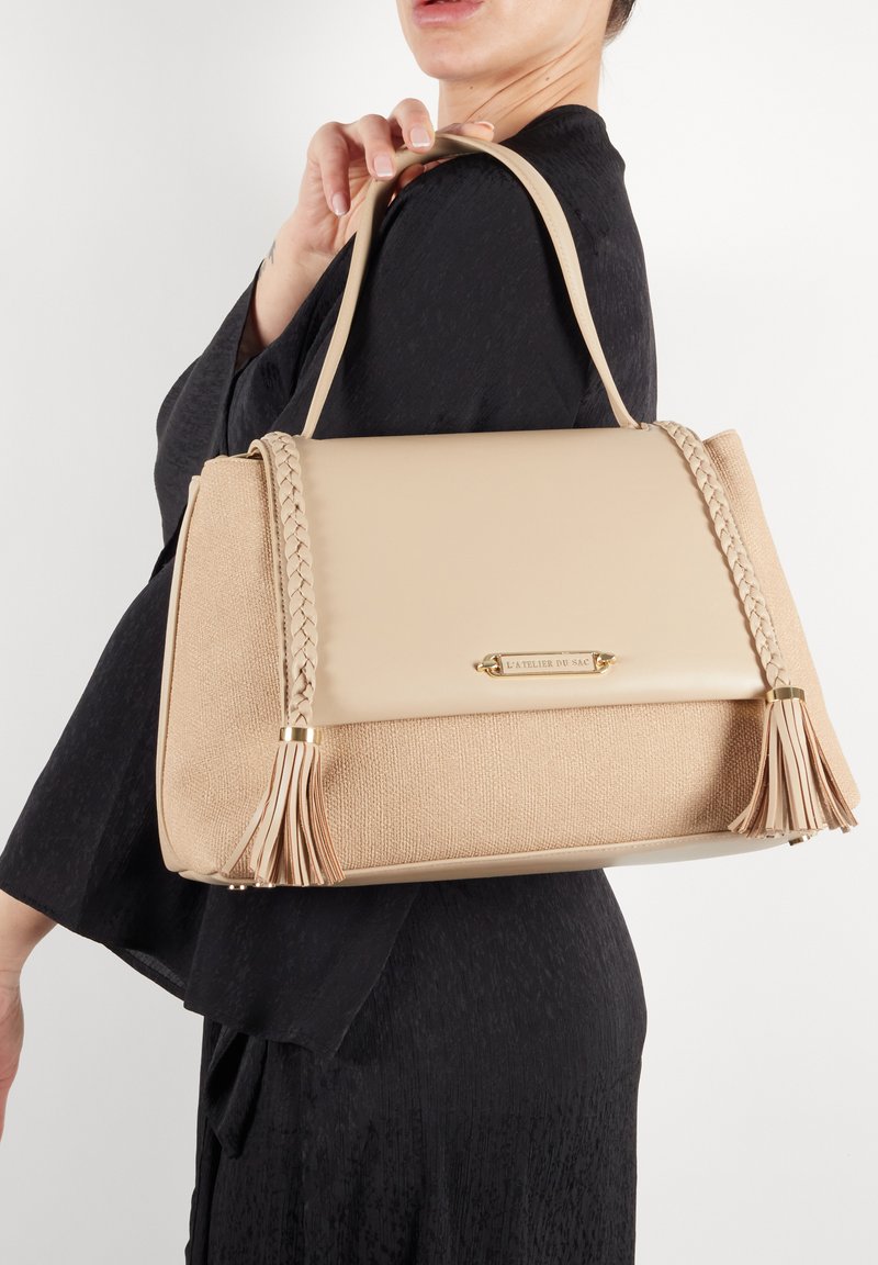 Beige håndtaske med tekstureret stof og glat læder, med flettede detaljer og frynser, guldbelægning og et struktureret design.