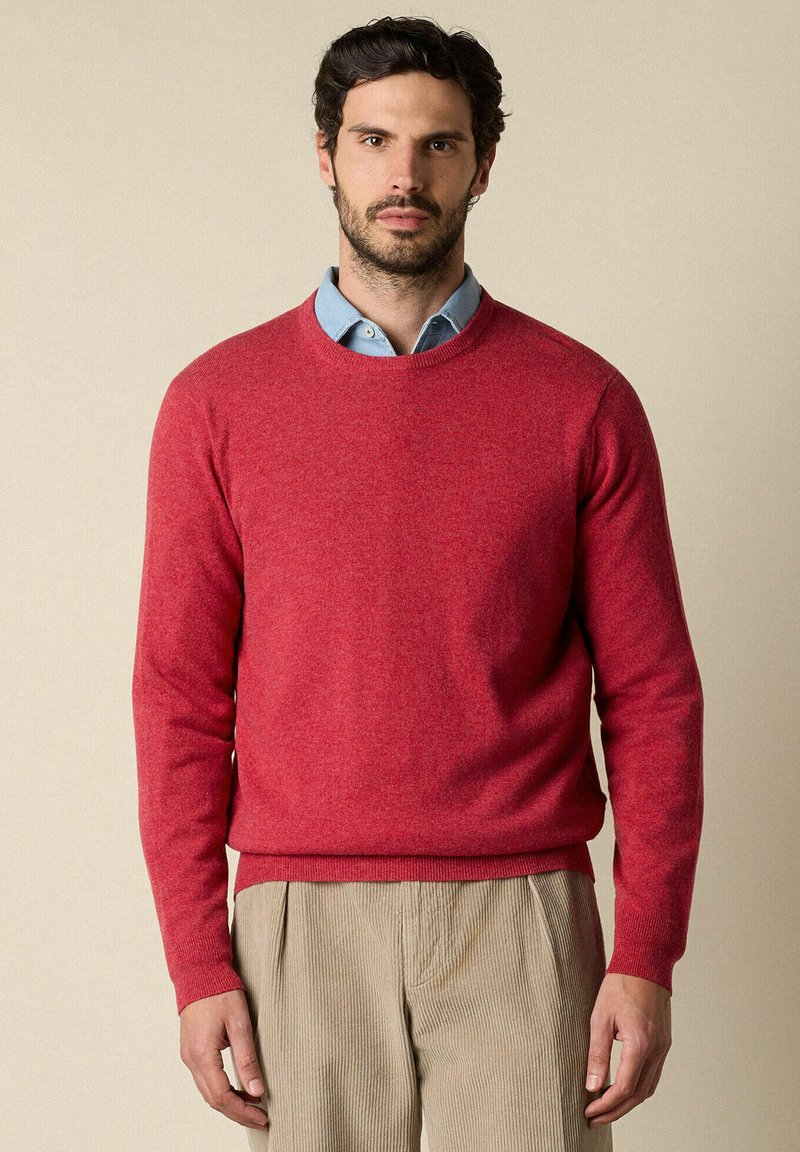 Roter Wollpullover mit rundem Ausschnitt, langen Ärmeln und gerippten Bündchen. Kombiniert mit einem blauen Hemd mit Kragen darunter.