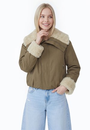 Jeune femme portant une veste verte avec un col et des poignets en peau lainée beige et un jean bleu clair, souriante et tenant son menton.