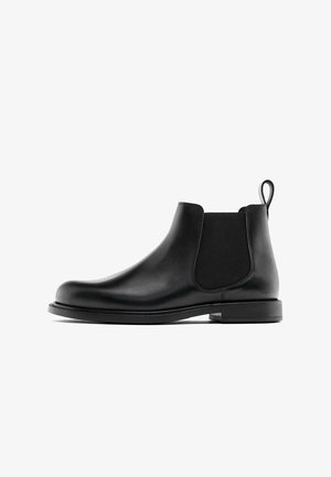 Bottine Chelsea en cuir noir avec des panneaux élastiques sur les côtés, une languette au talon et un petit talon empilé, sur fond blanc.