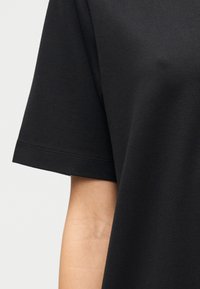 Μαύρο βαμβακερό μπλουζάκι με κοντά μανίκια, λεία υφή και κλασικό στρογγυλό γιακά, με ορατές ραφές στο στρίφωμα.