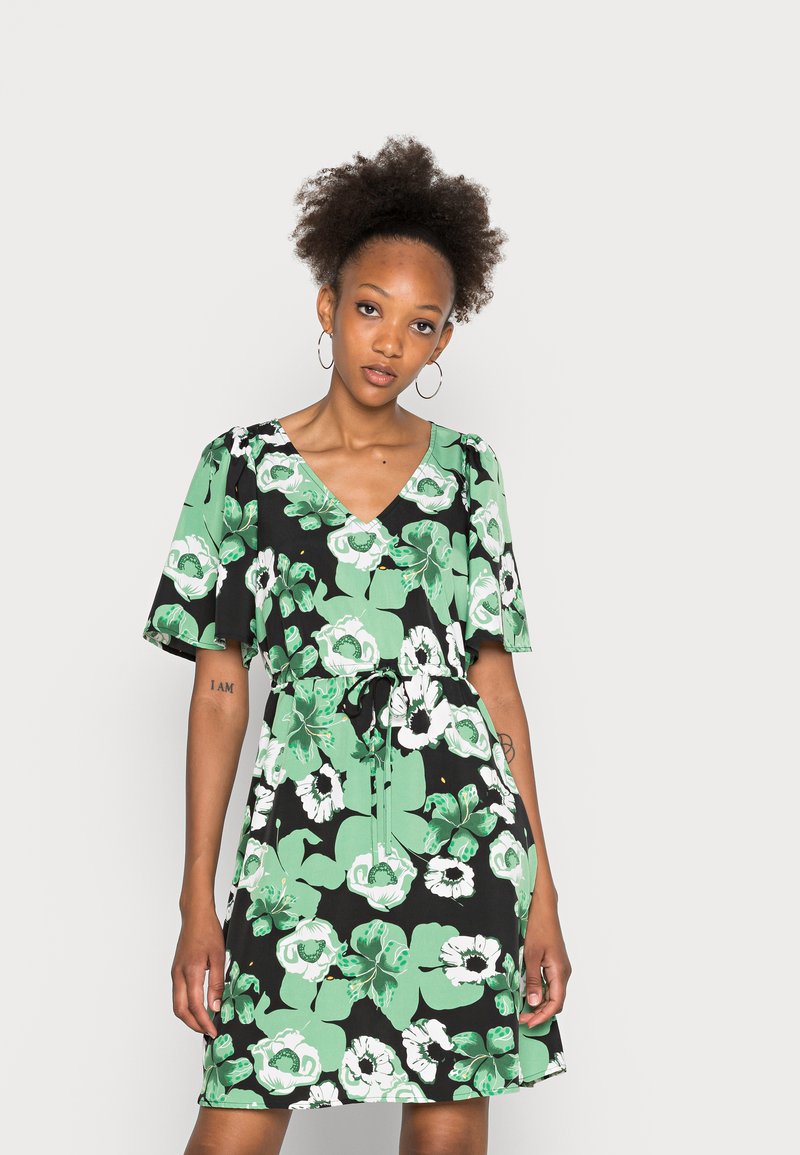 Vestido preto com padrão floral em verde e branco; mangas curtas, decote em V, cordão na cintura, criando um ajuste relaxado. Textura de tecido suave.