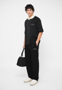 Jovem vestido com uma camisa preta de mangas curtas e abotoada da Armani Exchange e calças, segurando uma mala preta, com ténis brancos, num fundo branco.