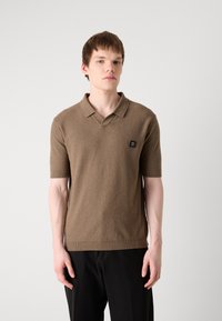 Brązowa koszulka polo z krótkim rękawem w teksturowanej tkaninie, z dekoltem w kształcie V i małą czarną naszywką z logo po lewej stronie klatki piersiowej. Noszona z ciemnymi spodniami.